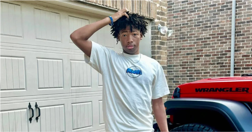 A young man with dreadlocks wearing a white t-shirt with a Pringles logo, standing in front of a red Jeep Wrangler.
