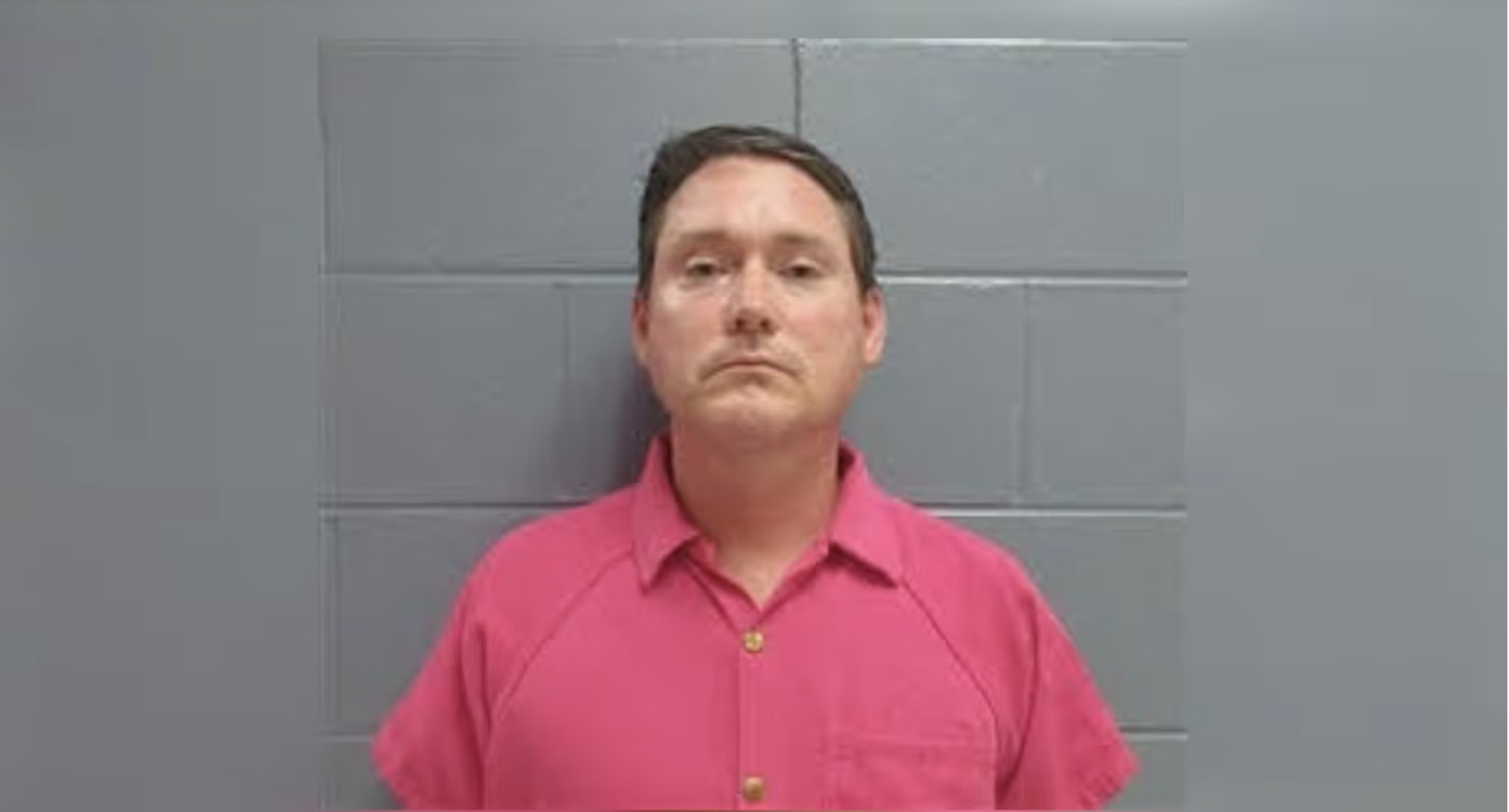 A man wearing a pink shirt standing in front of a gray tiled wall.