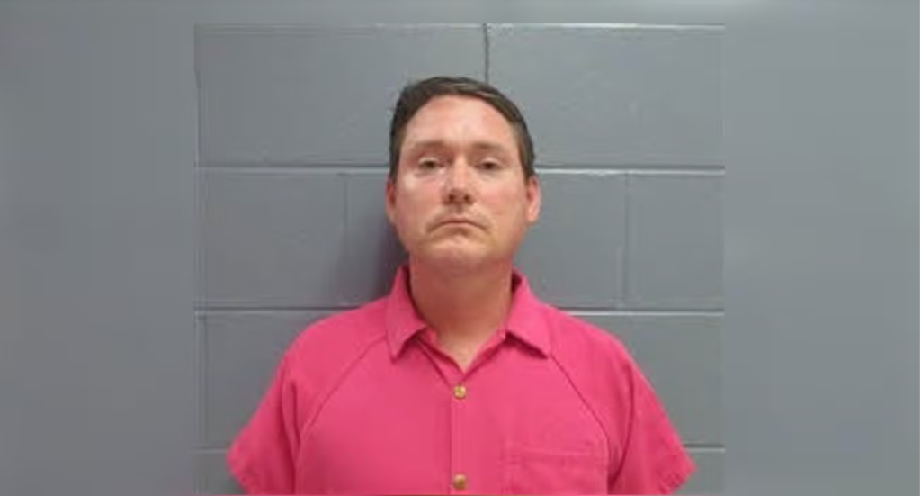 A man wearing a pink shirt standing in front of a gray tiled wall.