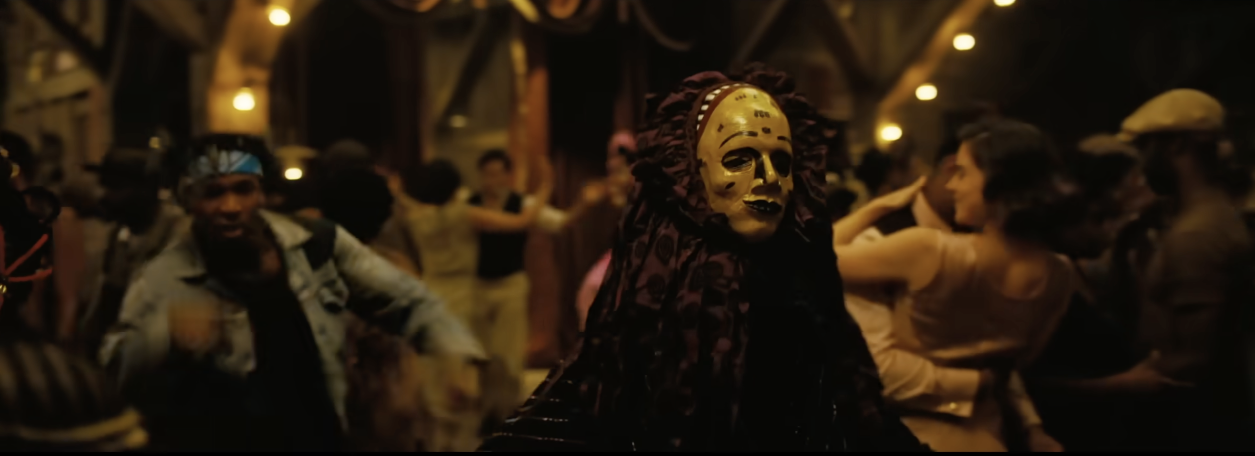 A group of people in costumes and masks, some wearing elaborate headdresses, gathered in a dimly lit setting with string lights.