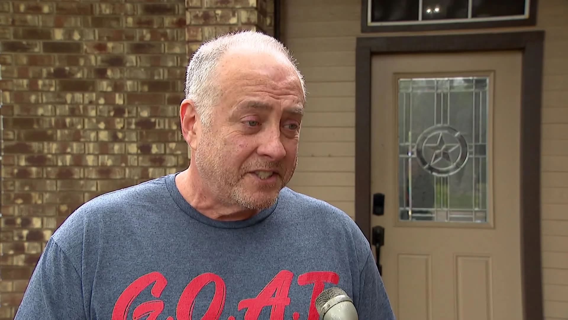 An older man with gray hair and a beard wearing a gray t-shirt with the text "G.O.A.T." standing in front of a brick building.