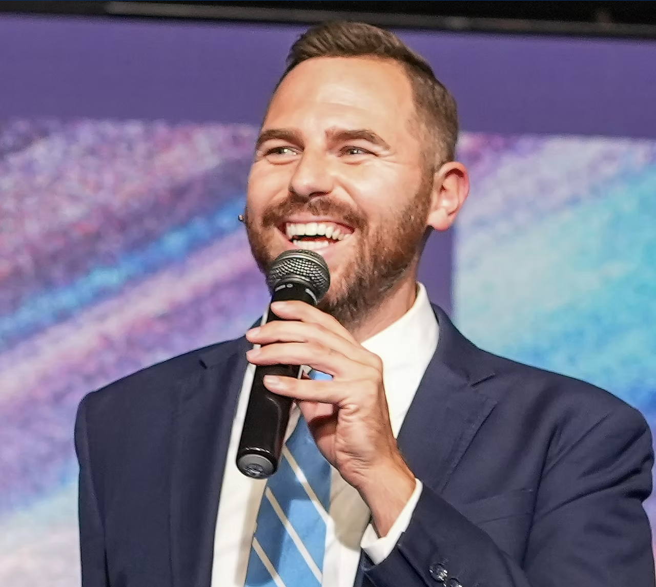A smiling man in a suit holding a microphone on a stage with a colorful background.