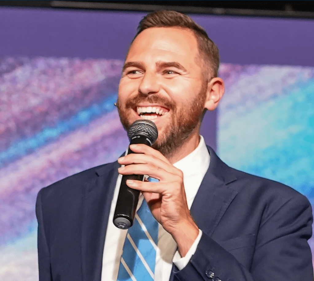 A smiling man in a suit holding a microphone on a stage with a colorful background.