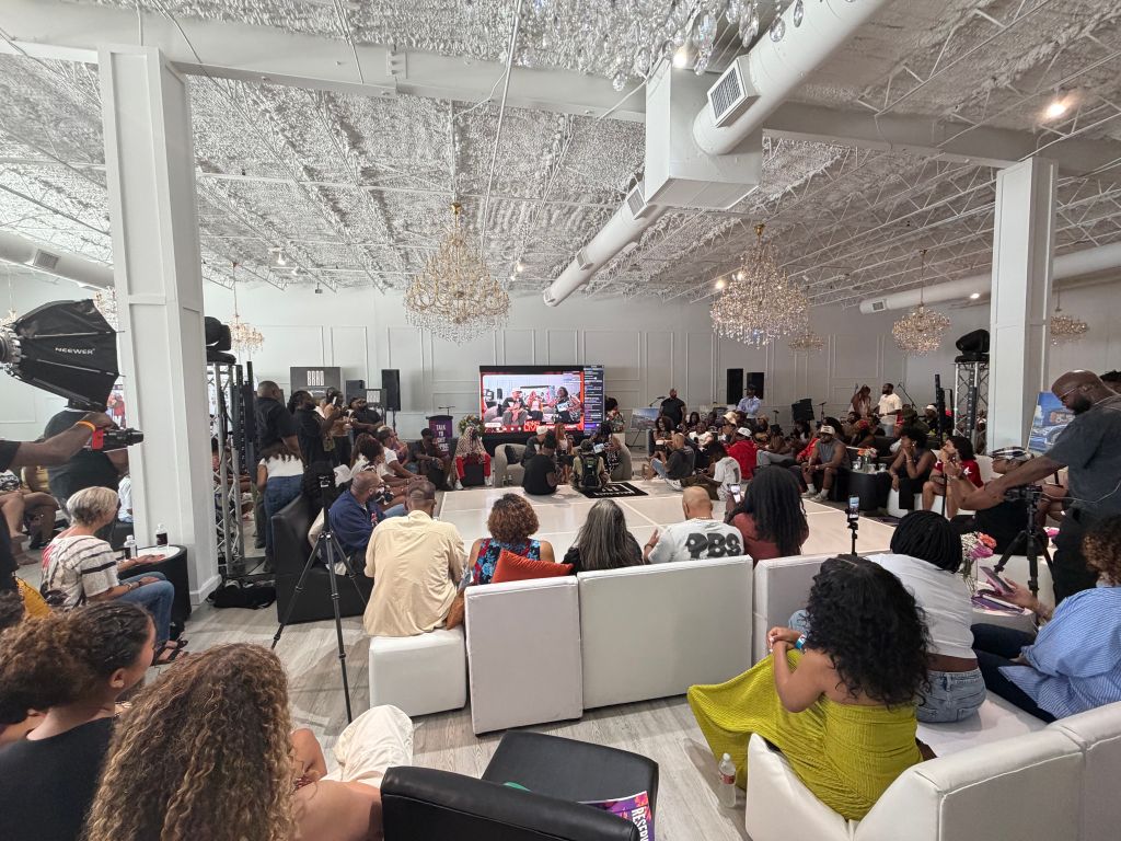 A crowded event space with people seated and standing, watching a presentation on a large screen. Several chandeliers and lighting fixtures are visible in the ceiling.