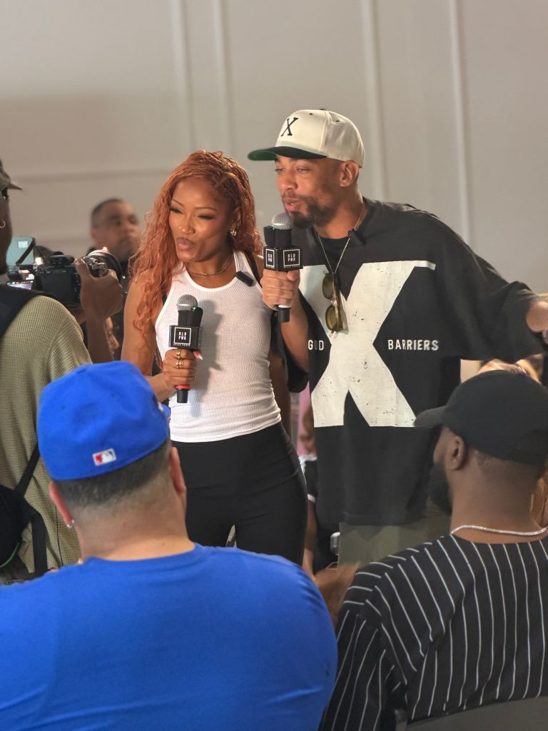 Two people, a woman with red hair and a man wearing a "Barriers" shirt, holding microphones and speaking to a crowd.