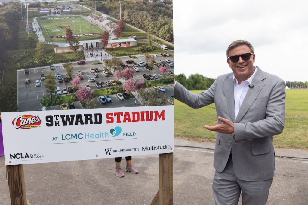 A man in a suit stands in front of a sign for Cane's 9th Ward Stadium at LCMC Health Field, with a large football stadium visible in the background.