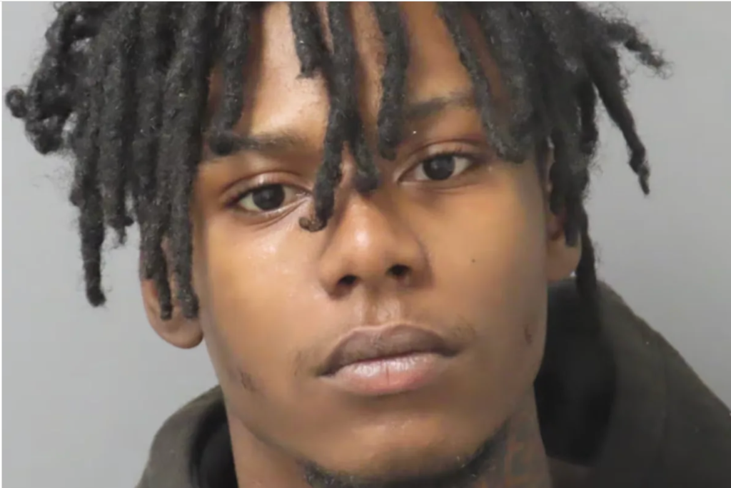 A close-up portrait of a young Black man with dreadlocks, looking directly at the camera with a serious expression.