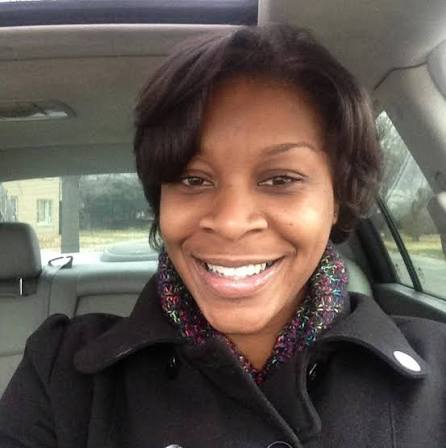 A smiling African American woman wearing a black coat and a colorful scarf in the front seat of a car.