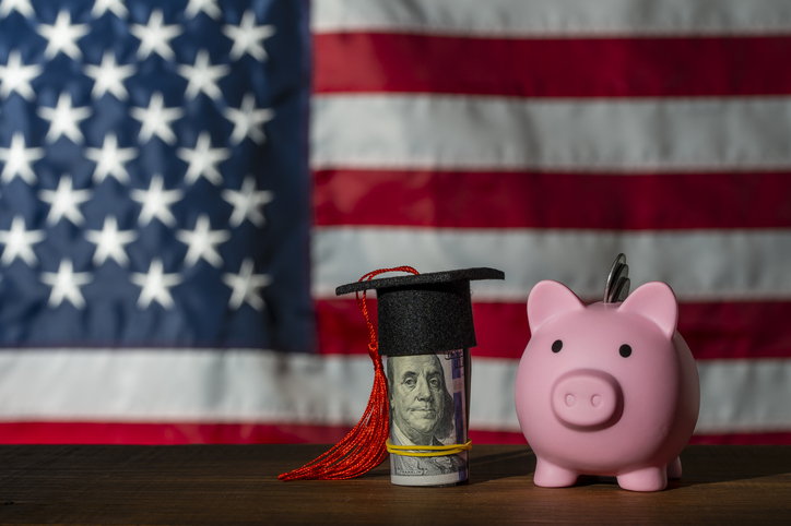 Graduation cap on dollar bill and piggy bank against US flag. A miniature graduation mortarboard hat on hundred dollar bill next to a pink piggy bank with the American flag blurred in the background
