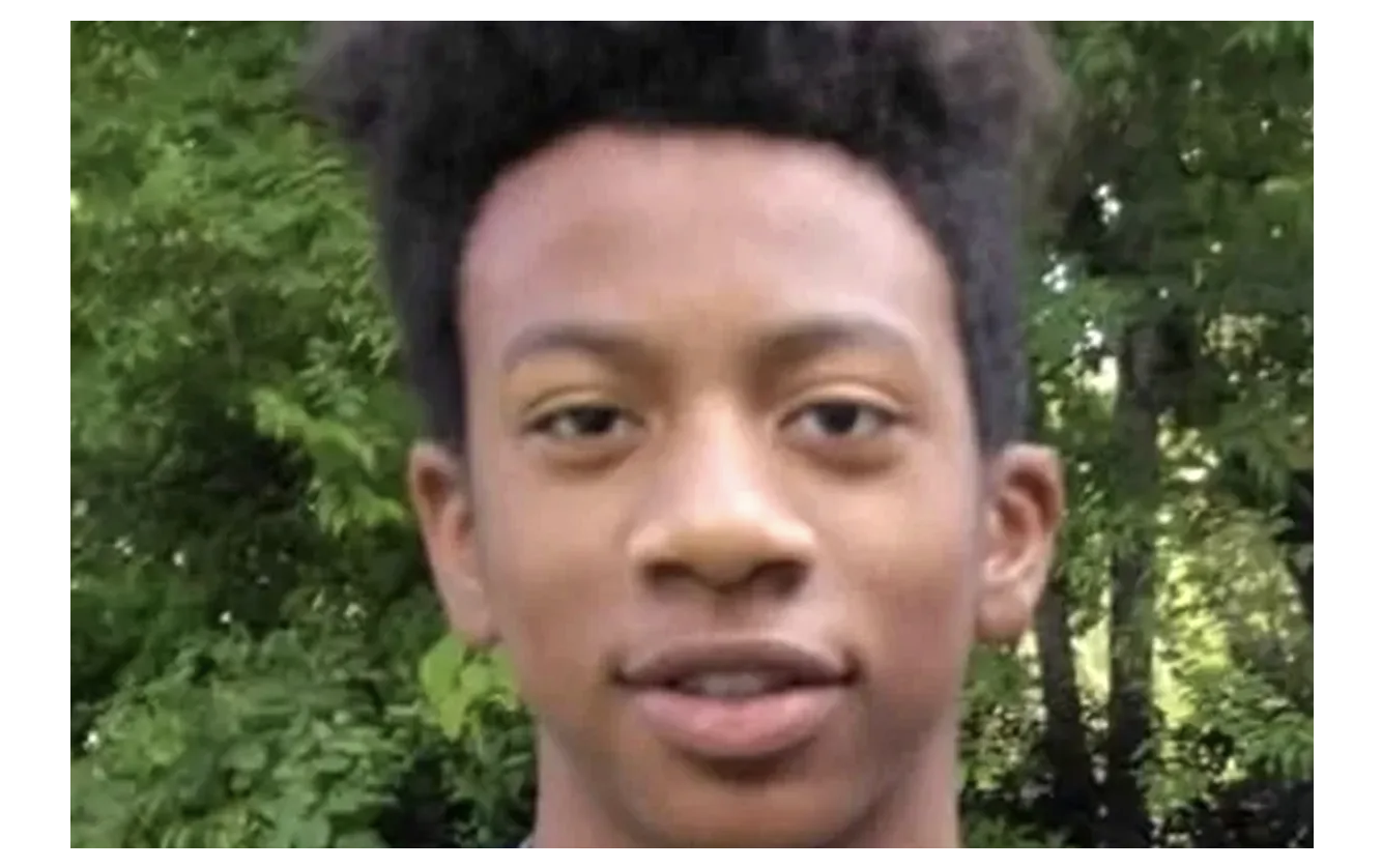 A young Black man with short curly hair looking directly at the camera against a background of green foliage.