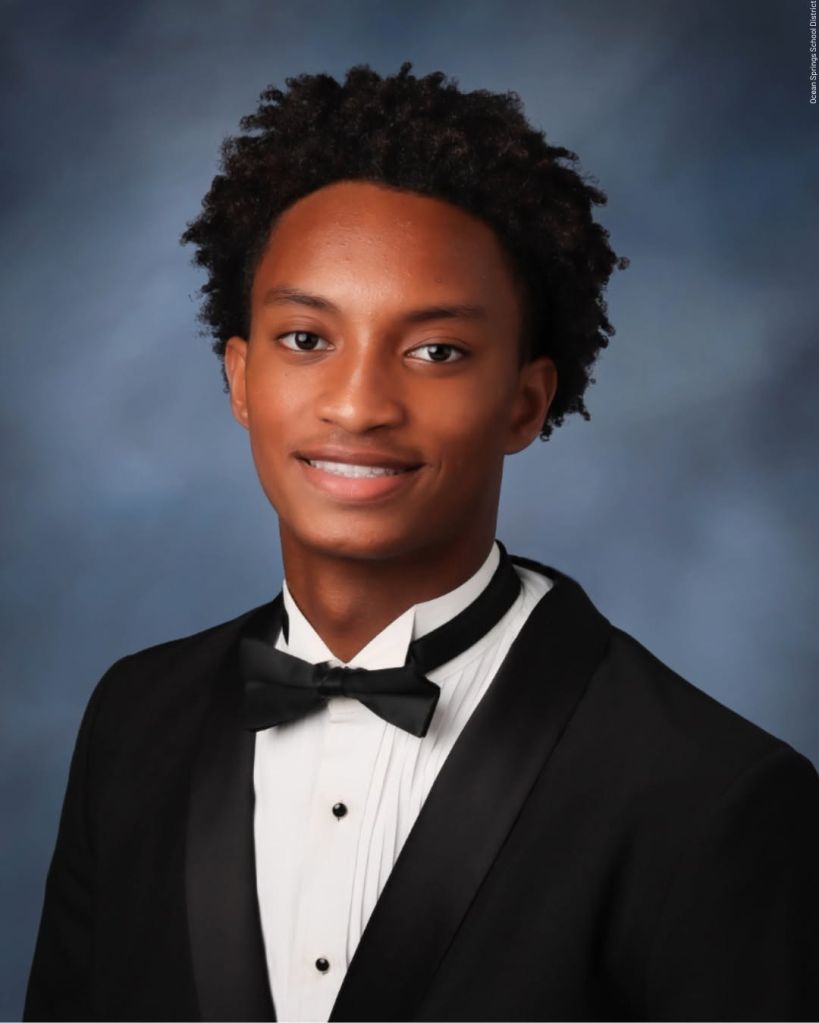 A young Black man wearing a tuxedo with a bow tie, smiling at the camera against a dark blue background.