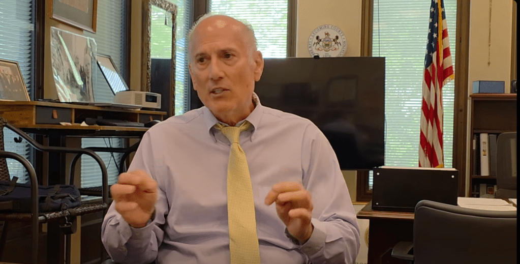 An older man in a light purple shirt and yellow tie gesturing while speaking in an office setting with an American flag visible.