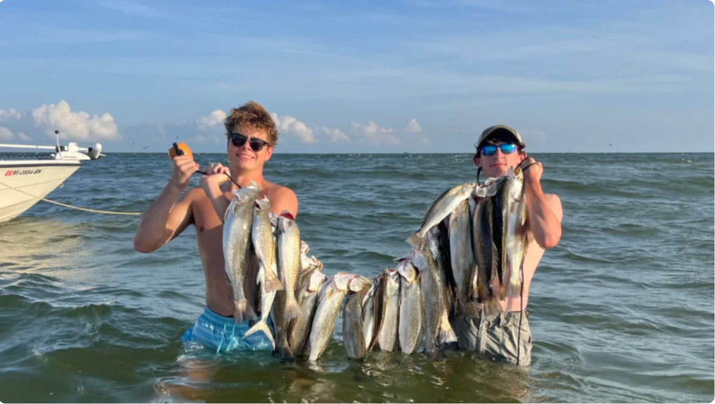 Two men holding up a large catch of fish in the ocean with a boat in the background.