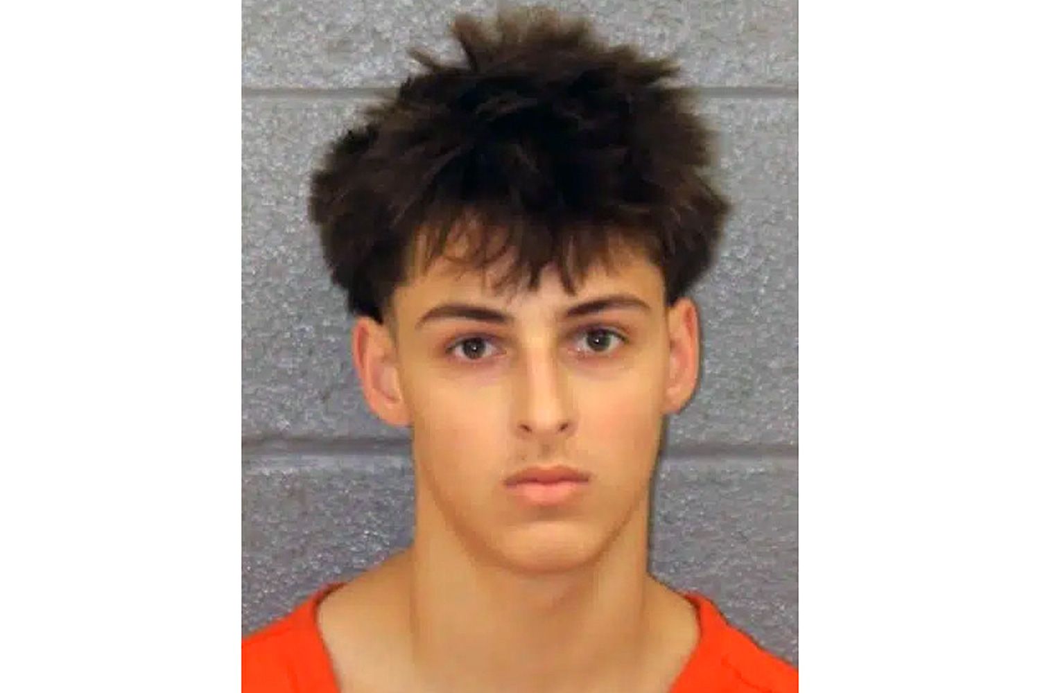 Mugshot of a young man with dark hair and a serious expression, wearing an orange shirt against a gray concrete wall.