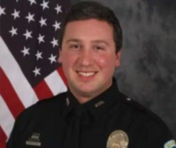 A smiling man wearing a police uniform and standing in front of an American flag.