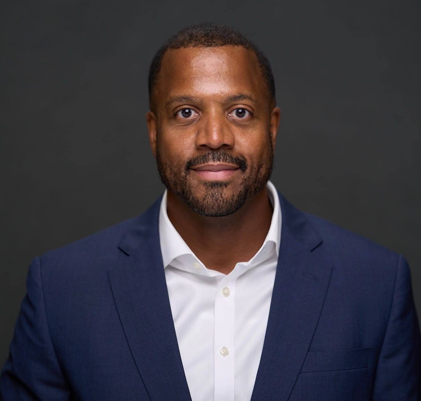 A headshot of a middle-aged Black man with a beard, wearing a navy blue suit and white shirt, looking directly at the camera against a dark background.