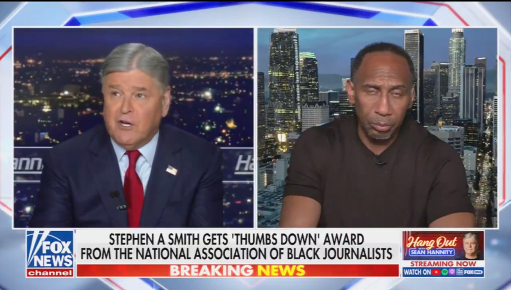 Two news anchors, one on the left wearing a red tie and the other on the right wearing a brown shirt, discussing a news story on the screen behind them showing a city skyline at night.