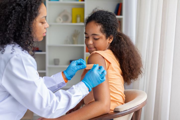 Black female doctor applying adhesive bandage to girls upper arm after vaccination