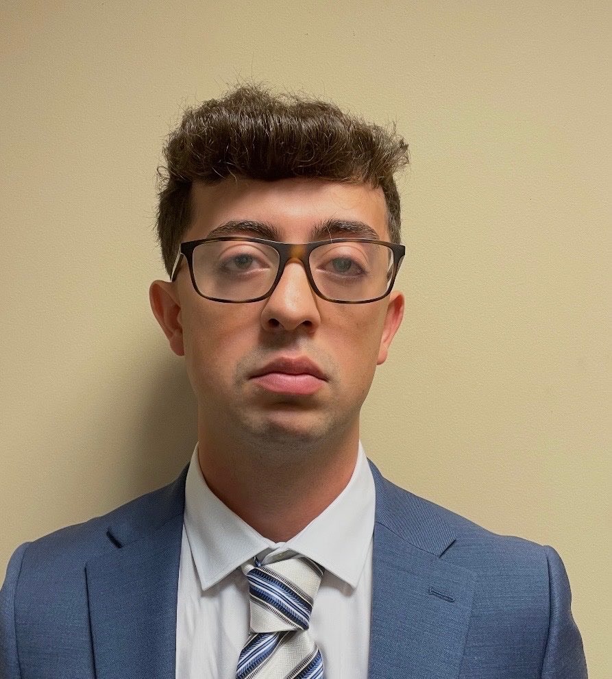 A man wearing glasses and a suit, with a serious expression on his face, standing against a plain background.