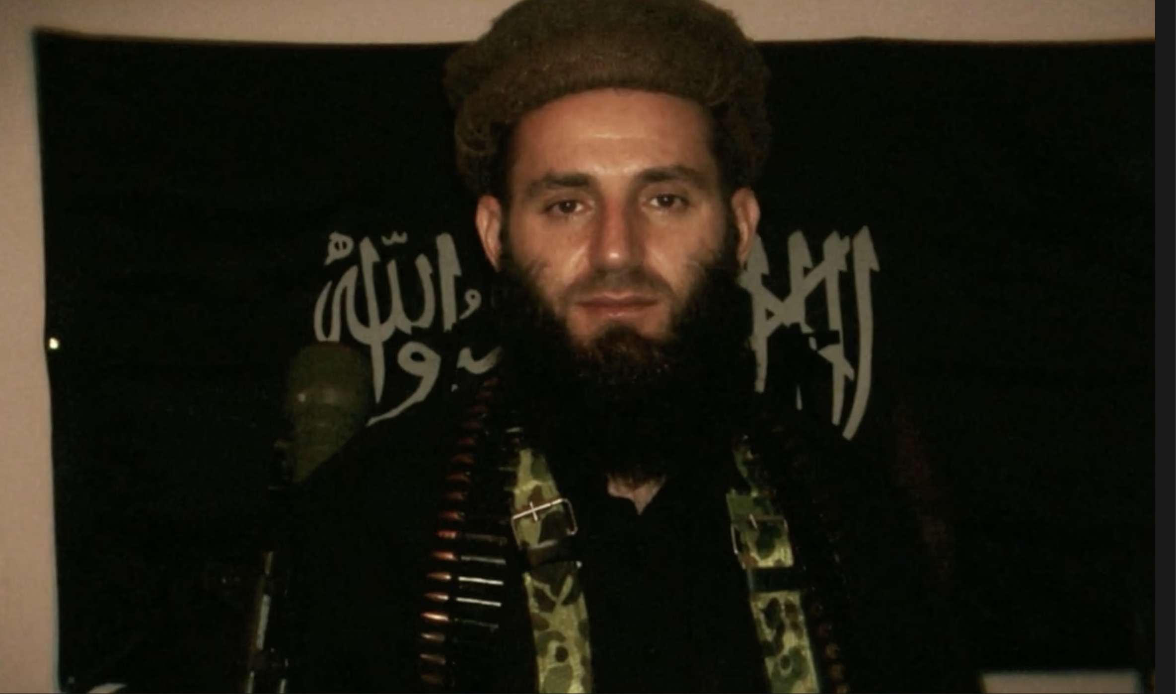A bearded man wearing a camouflage headband and clothing, holding a firearm in front of a black flag with Arabic text.