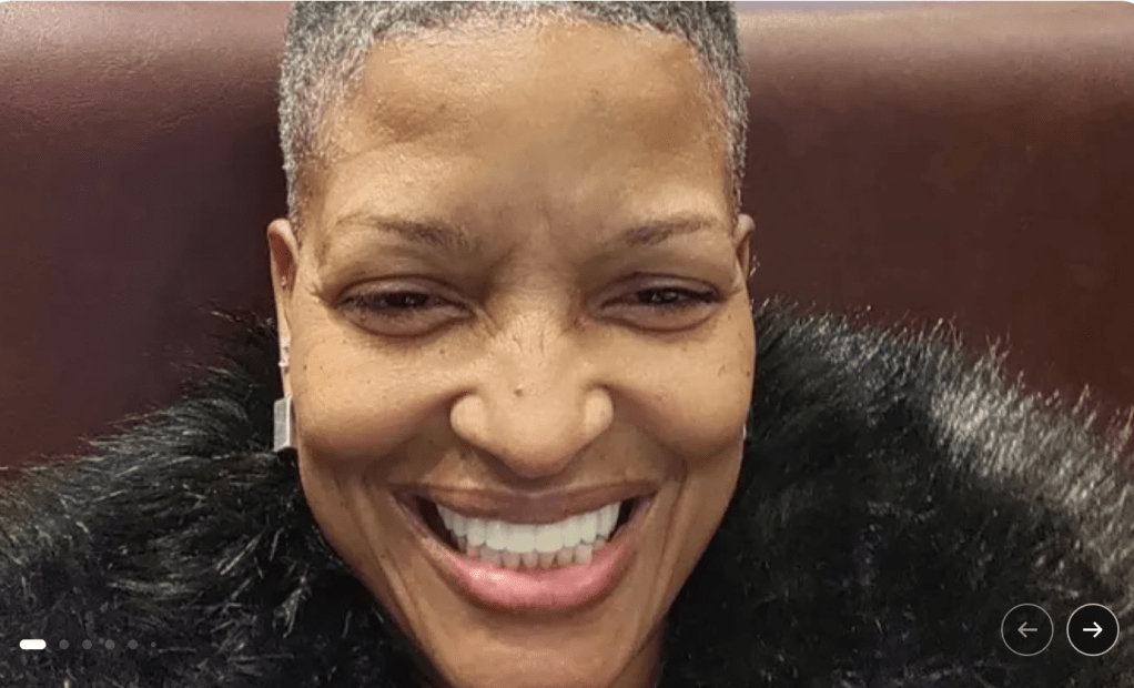 A smiling woman with short gray hair wearing a black feathered coat.