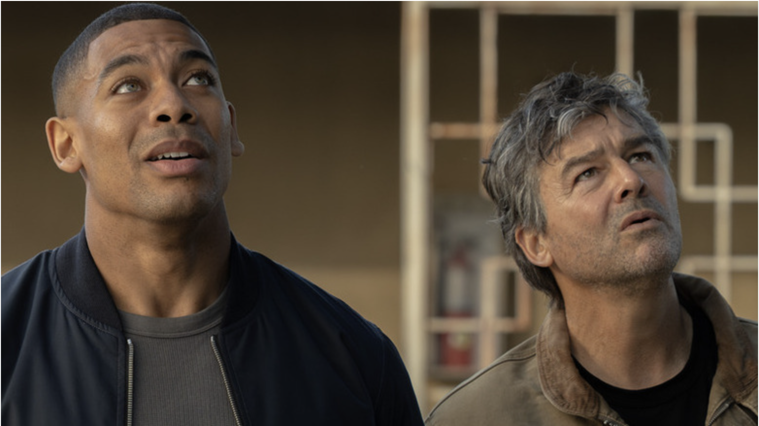 Two men, one younger and one older, looking up with concerned expressions in a dimly lit room.