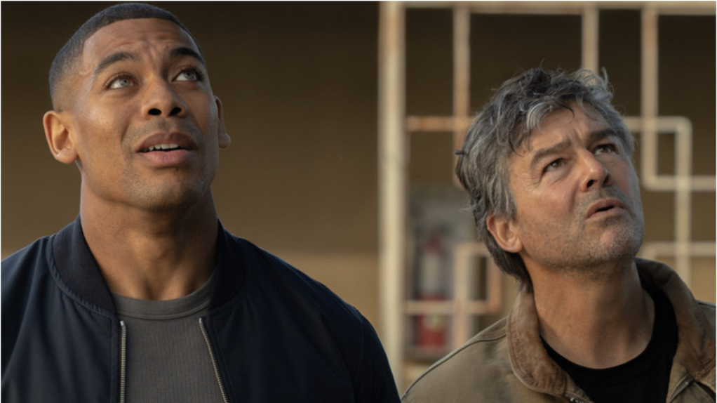 Two men, one younger and one older, looking up with concerned expressions in a dimly lit room.