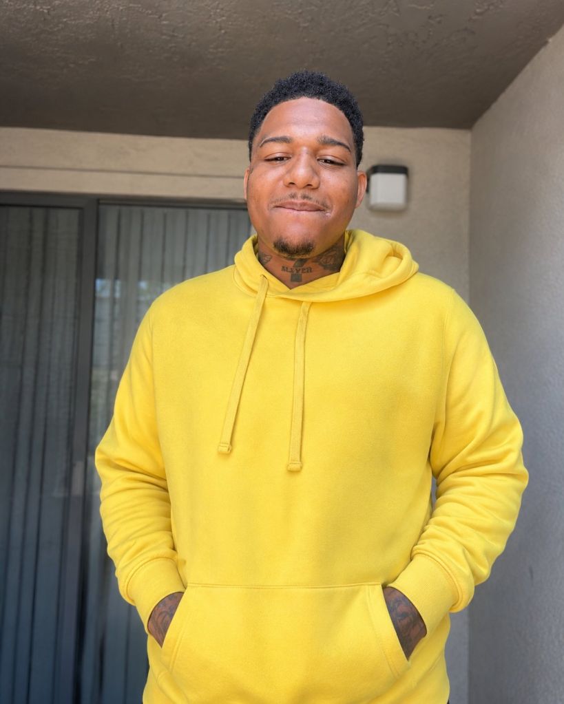 A young man wearing a yellow hooded sweatshirt with his hands in the pockets, standing in front of a building.