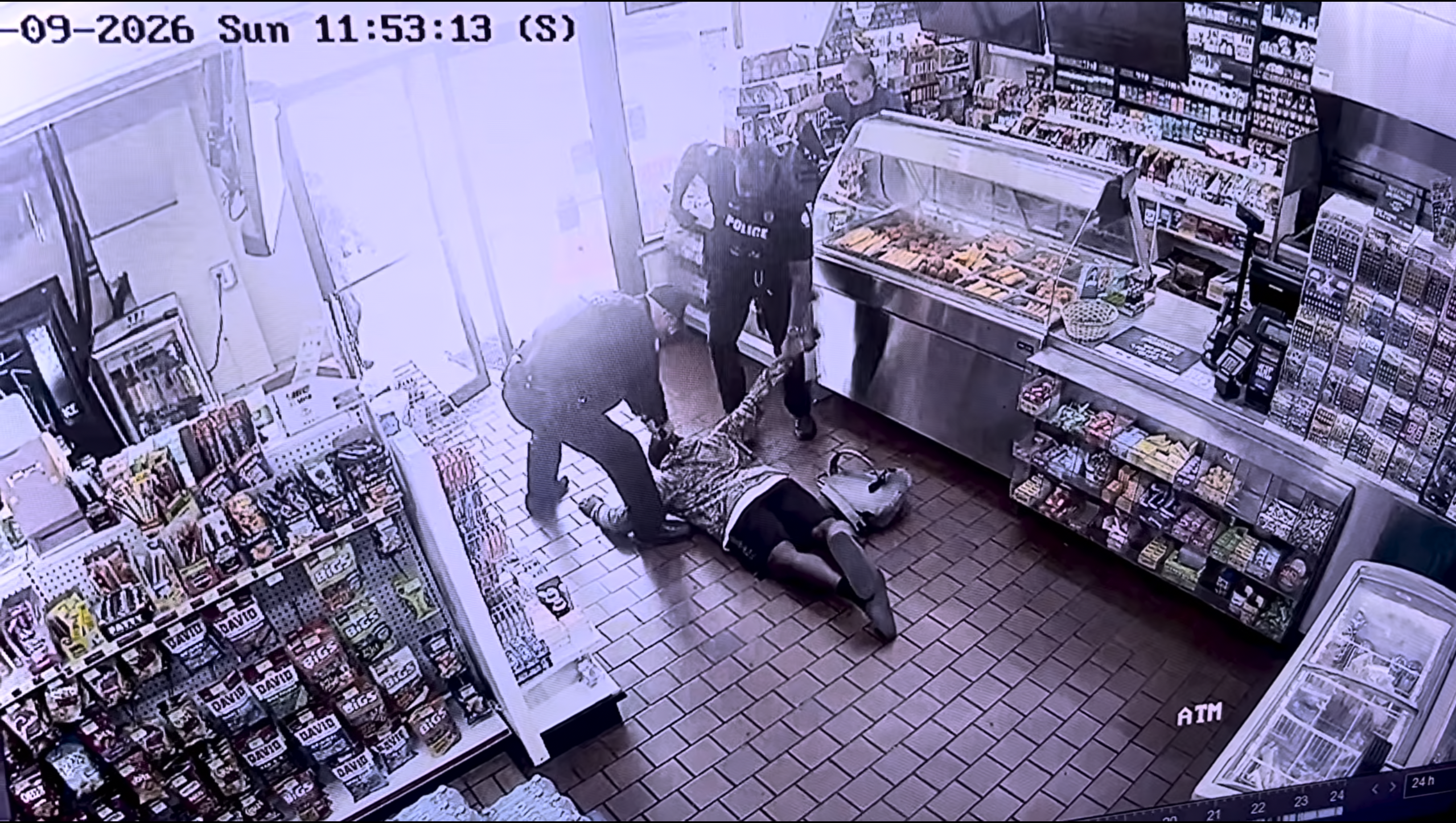 A security camera captures a person lying on the floor of a convenience store, with another person standing over them.